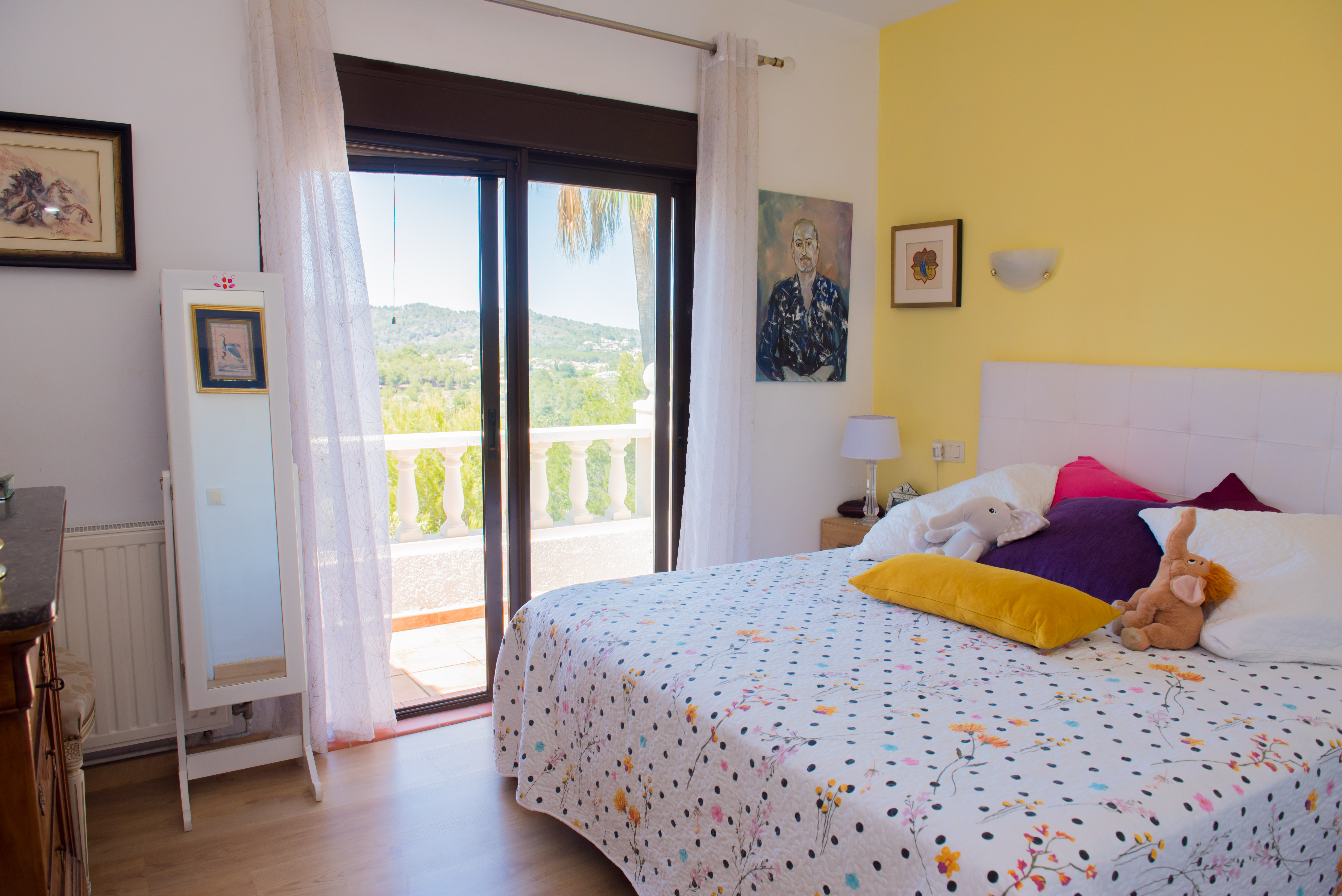 Bright bedroom with balcony access. Features a yellow accent wall and patterned bedding.