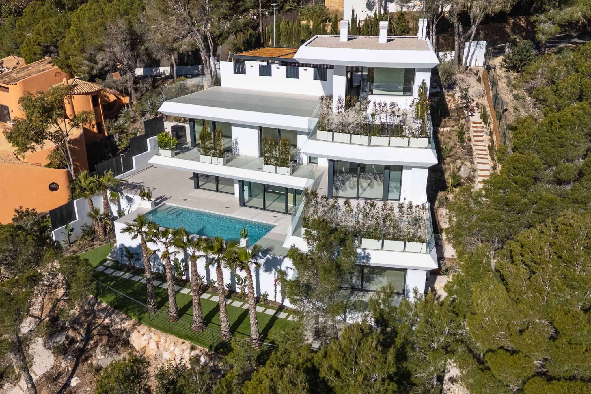Aerial view of a modern multi-level white villa nestled into a green hillside. Features a large private swimming pool, extensive terraces with glass railings, and palm trees, surrounded by Mediterranean forest.