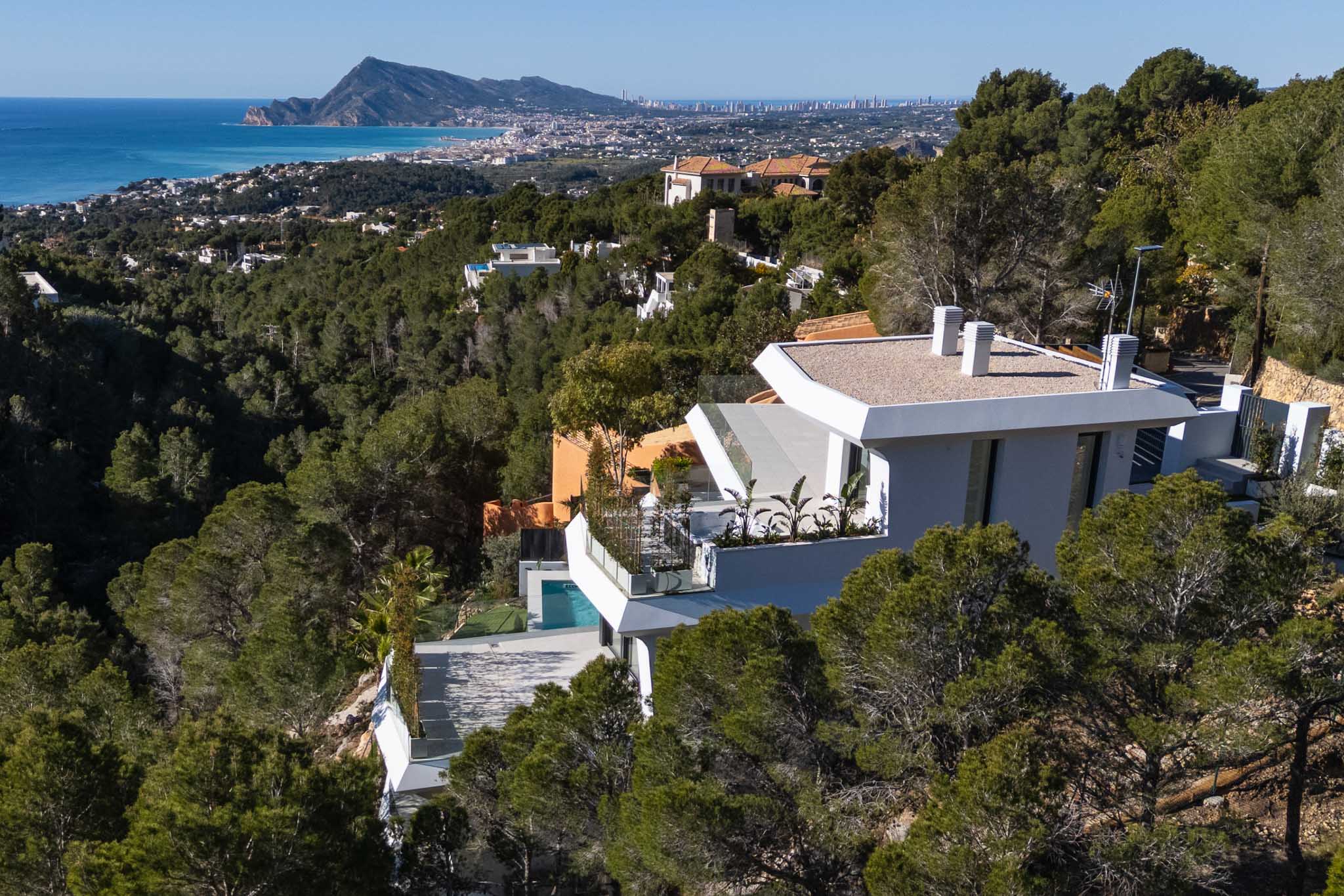 Aerial view of a modern white villa cascading down a pine-covered hillside, offering expansive panoramic views of the Mediterranean Sea and Altea's coastline. A private pool is visible on a lower terrace.