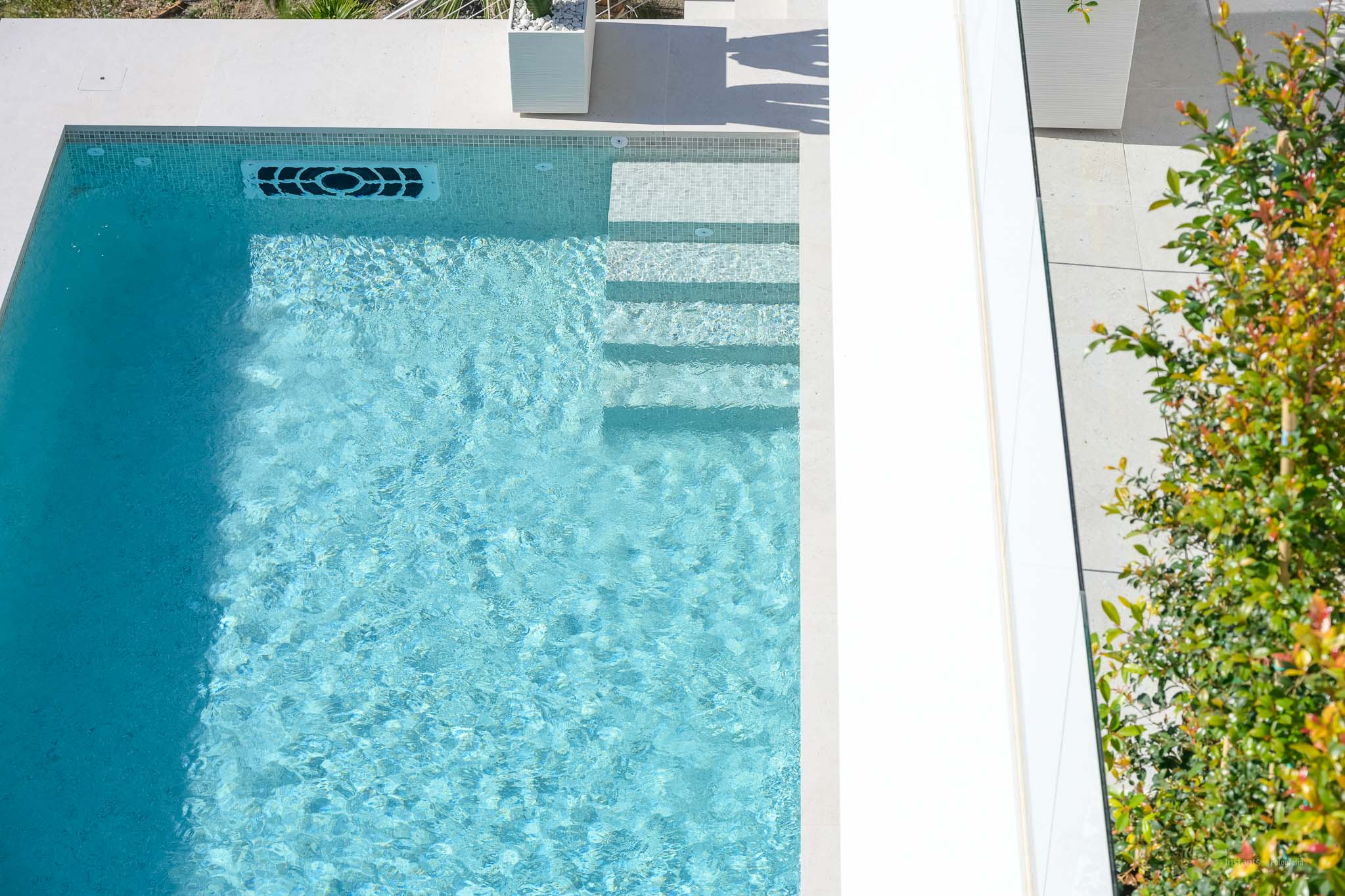 Aerial view of the light blue mosaic tiled pool with integrated steps, featuring a modern water return system and surrounding light-colored paving.