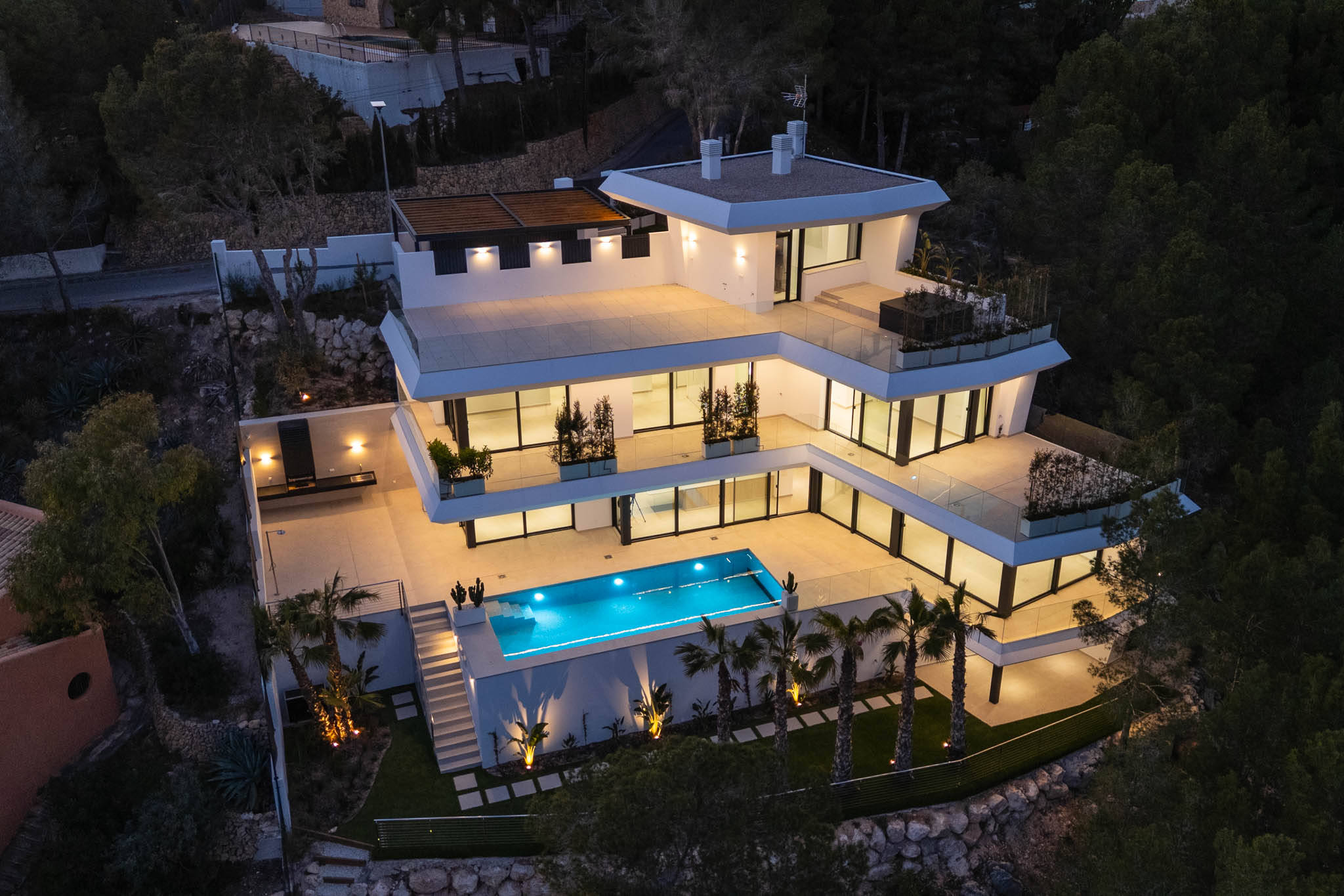 Aerial view of the modern multi-level villa at dusk, showcasing its illuminated terraces, bright blue swimming pool, and an outdoor kitchen area with elegant lighting amidst lush greenery.