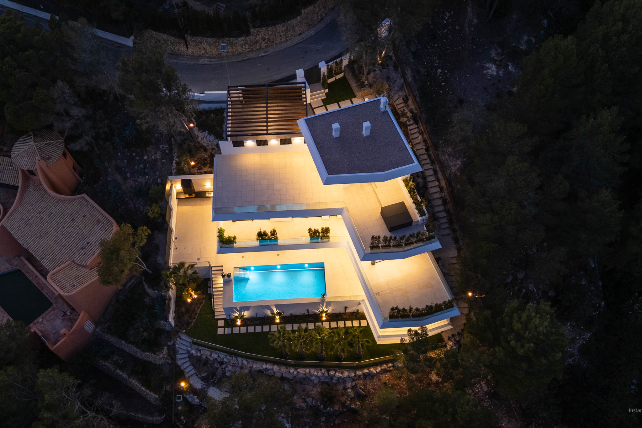 Aerial view of the modern villa at night, showcasing its brightly lit terraces and the inviting, glowing blue swimming pool. Multiple outdoor living areas are visible, set against the dark, natural hillside.