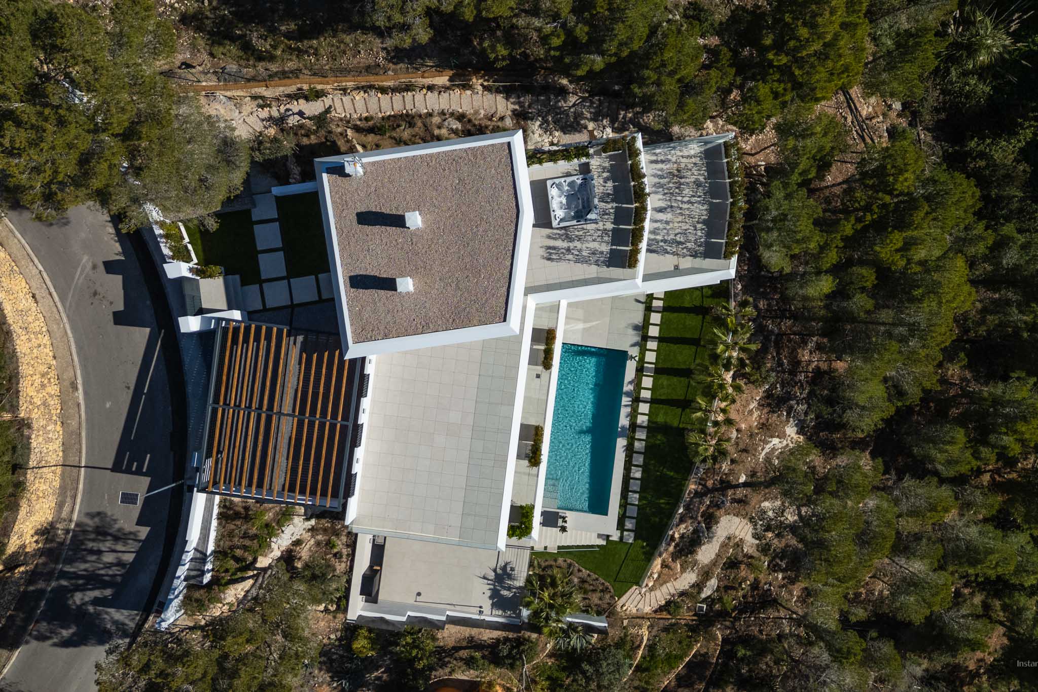 Aerial view showcasing the modern villa's architecture, including a rectangular swimming pool, expansive paved terraces, and an outdoor spa, all nestled within natural Mediterranean greenery.