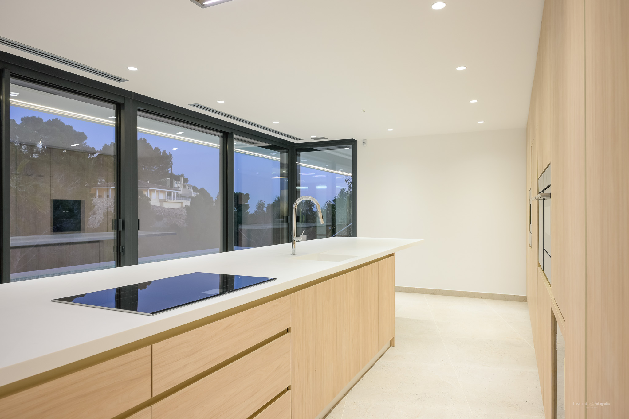 Contemporary kitchen featuring a large central island with a white countertop, integrated induction hob, and chrome faucet. Light wood cabinetry and built-in ovens line the wall, complemented by expansive windows.