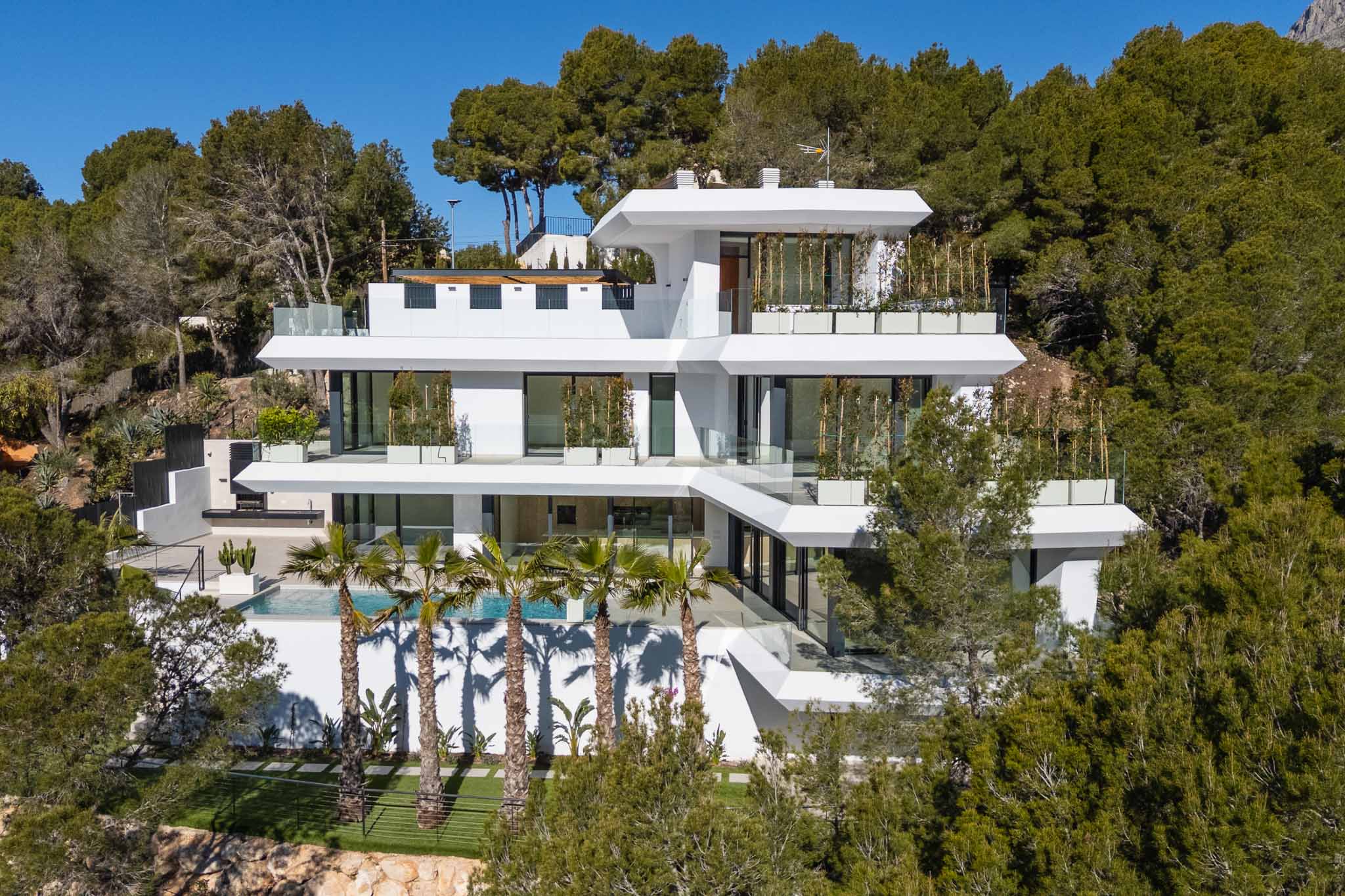 Contemporary multi-level villa featuring a pristine white facade, expansive glass terraces, a swimming pool with palm trees, and an outdoor kitchen, integrated into a lush green hillside.