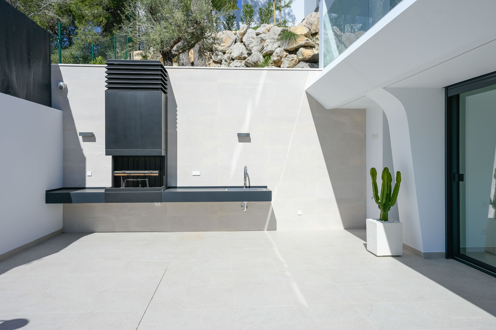 Contemporary outdoor BBQ area featuring an integrated grill and sink, set on light-toned tiled flooring. A potted cactus and natural rock landscaping enhance the space.