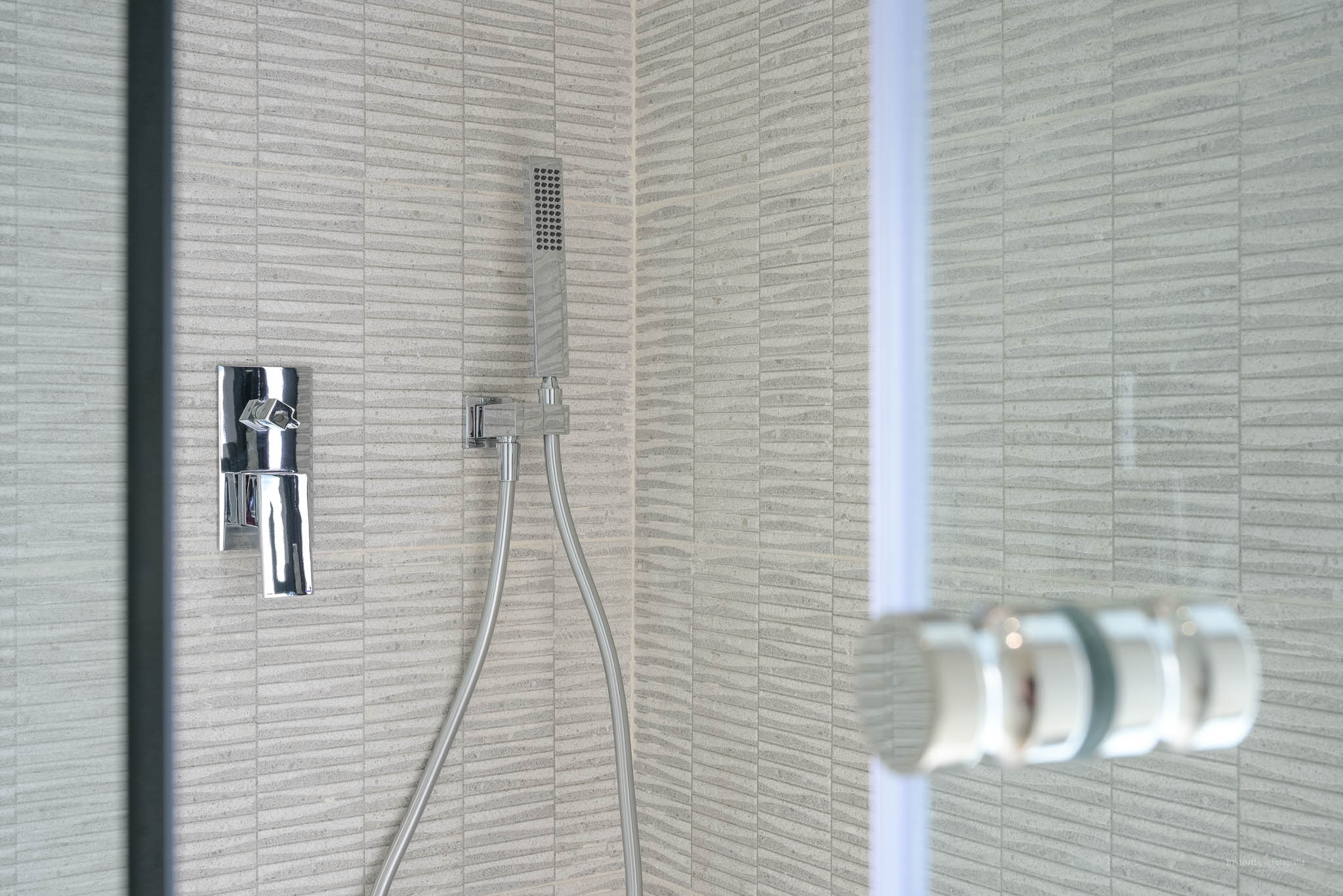 Detail of a contemporary shower featuring grey textured rectangular tiles, a sleek chrome mixer tap, and a flexible handheld showerhead. A glass screen is partially visible.