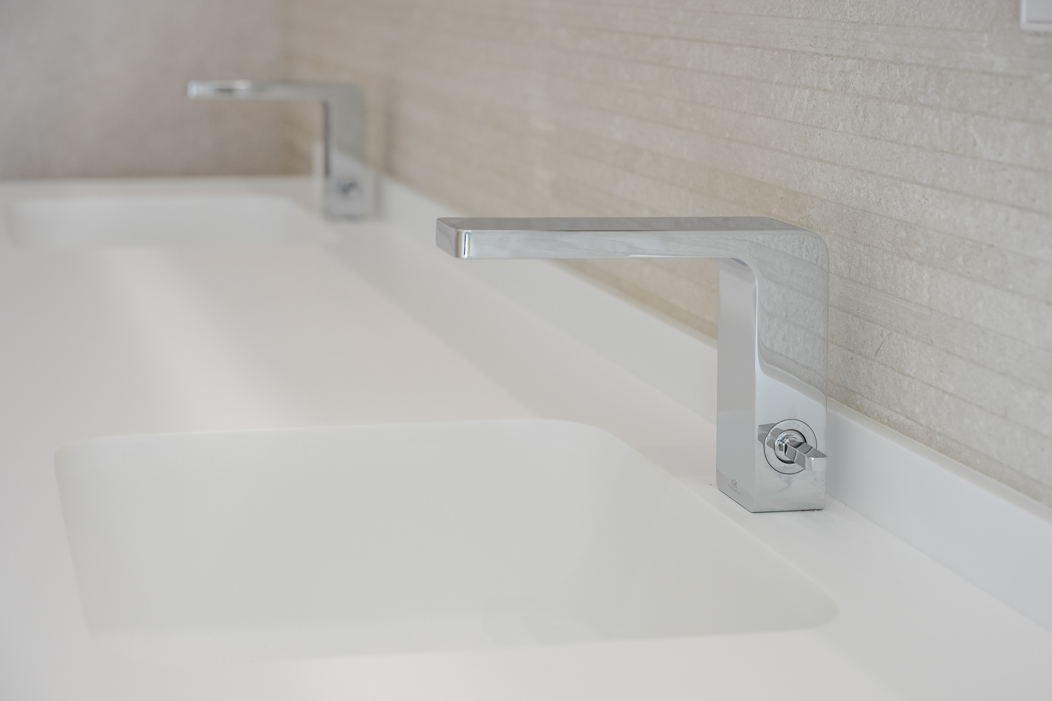 Double vanity featuring sleek chrome faucets and integrated white sinks. The background wall is clad with light, horizontally textured tiles.