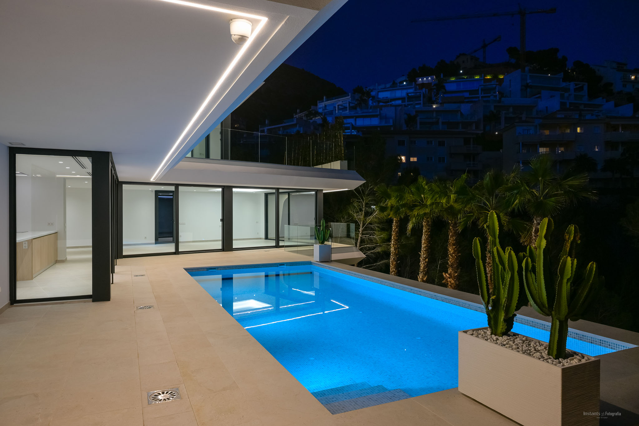 Illuminated swimming pool at night, featuring vibrant blue underwater lighting and a spacious tiled terrace. Modern linear ceiling lights enhance the sleek architecture, overlooking a hillside with distant buildings.