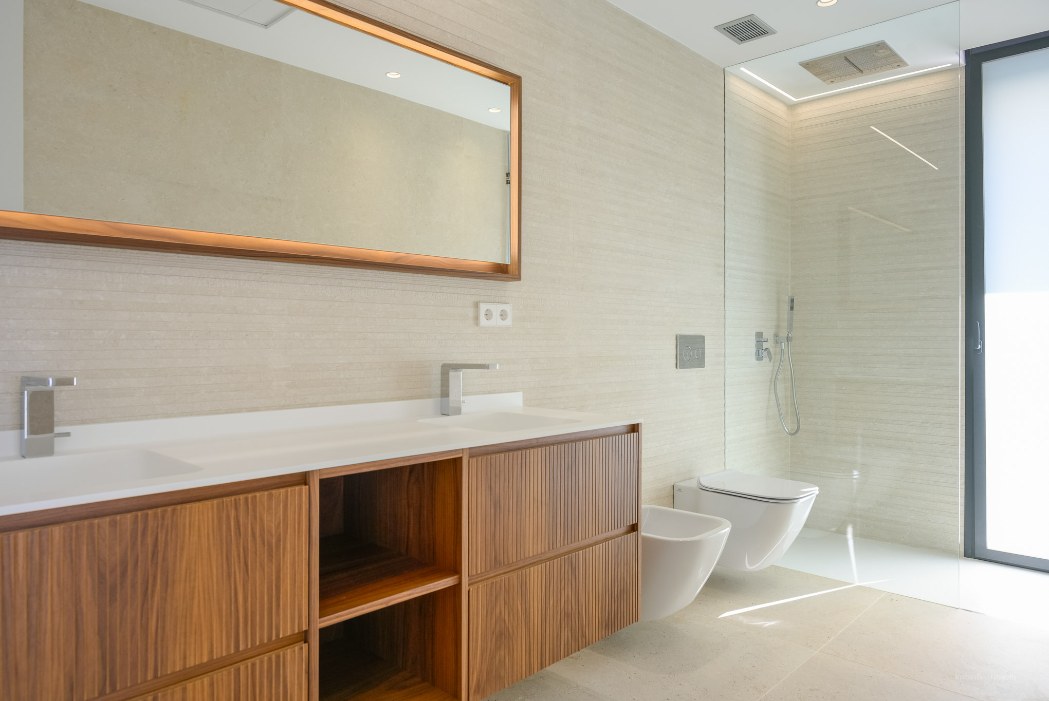 Modern bathroom featuring a double vanity with wooden cabinets, textured wall tiles, and an illuminated mirror. Includes a walk-in shower, wall-mounted toilet, and bidet.
