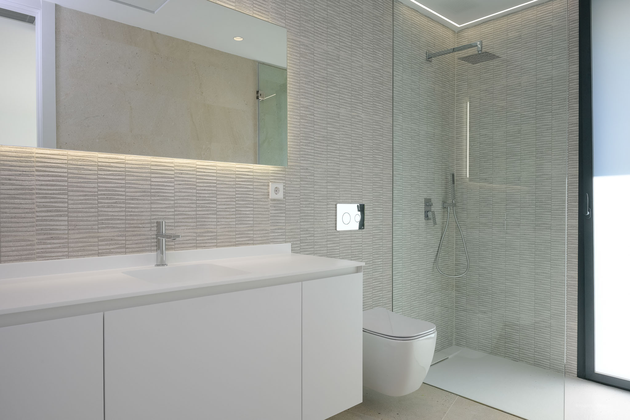 Modern bathroom featuring a sleek white vanity with integrated sink, backlit mirror, and textured grey wall tiles. A wall-mounted toilet and spacious walk-in shower with glass partition.