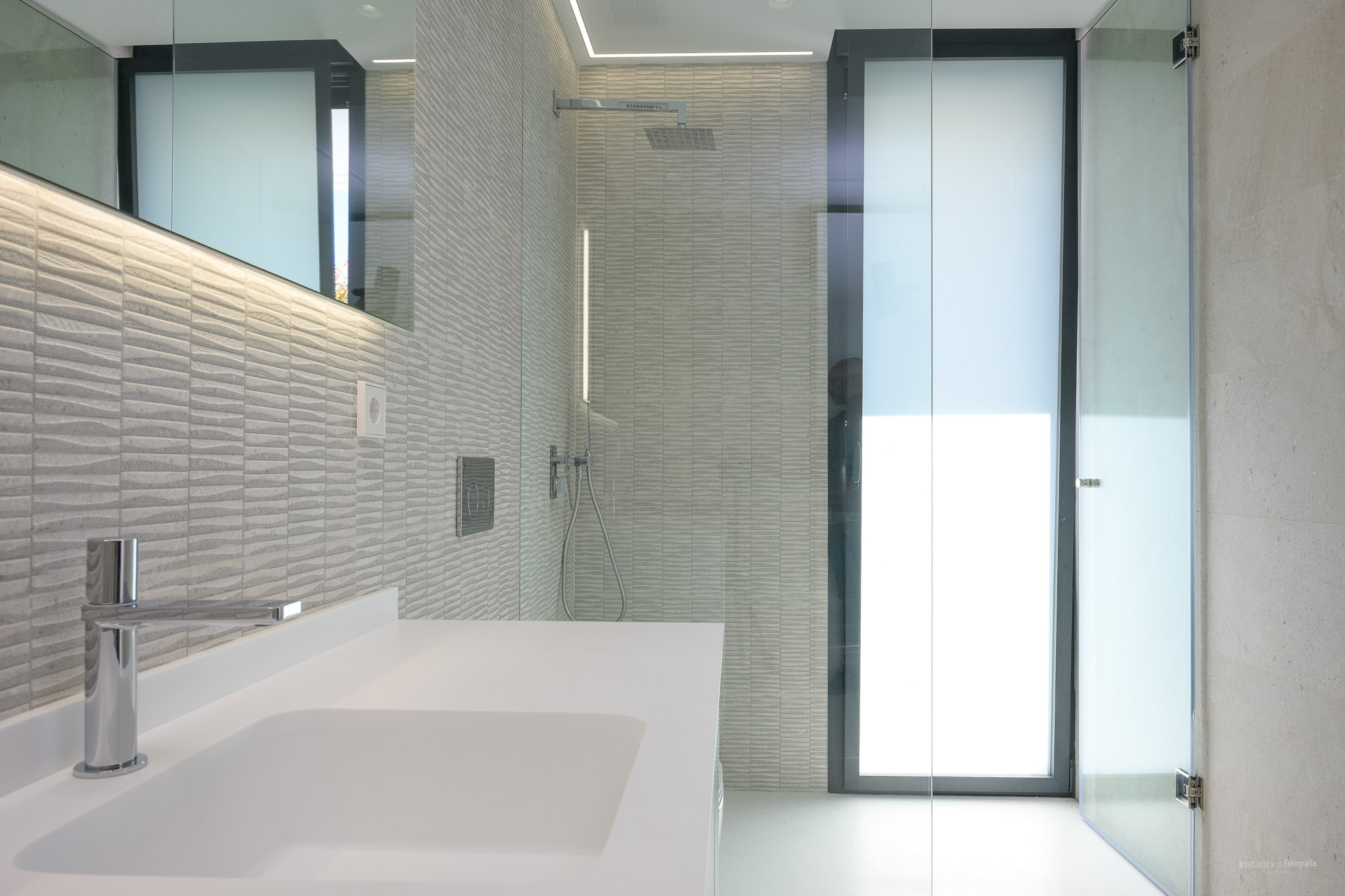 Modern bathroom featuring a white basin, chrome faucet, and textured grey wall tiles. Includes a glass-enclosed shower with a rainfall head and LED-lit mirror.