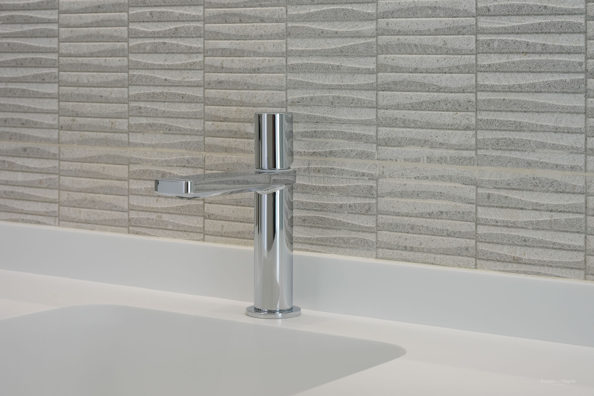 Modern chrome faucet on a white integrated sink countertop, complemented by textured light-colored wall tiles featuring a subtle wave pattern.