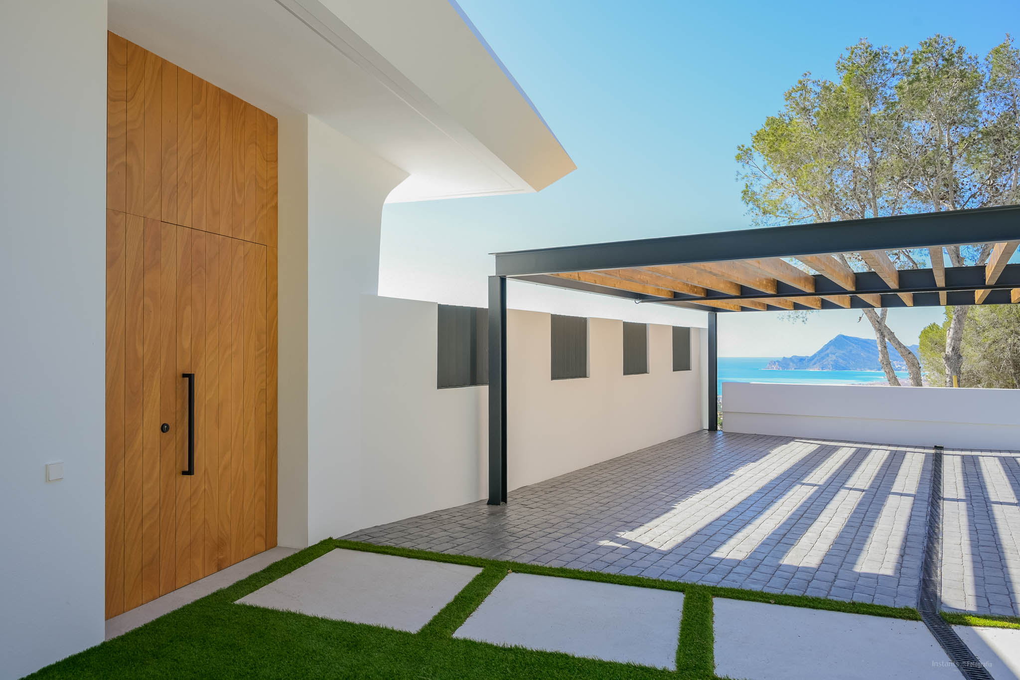 Modern entrance featuring a tall wooden door, artificial grass path, and a paved parking area with a metal pergola. Distant Mediterranean sea and mountain views are visible.