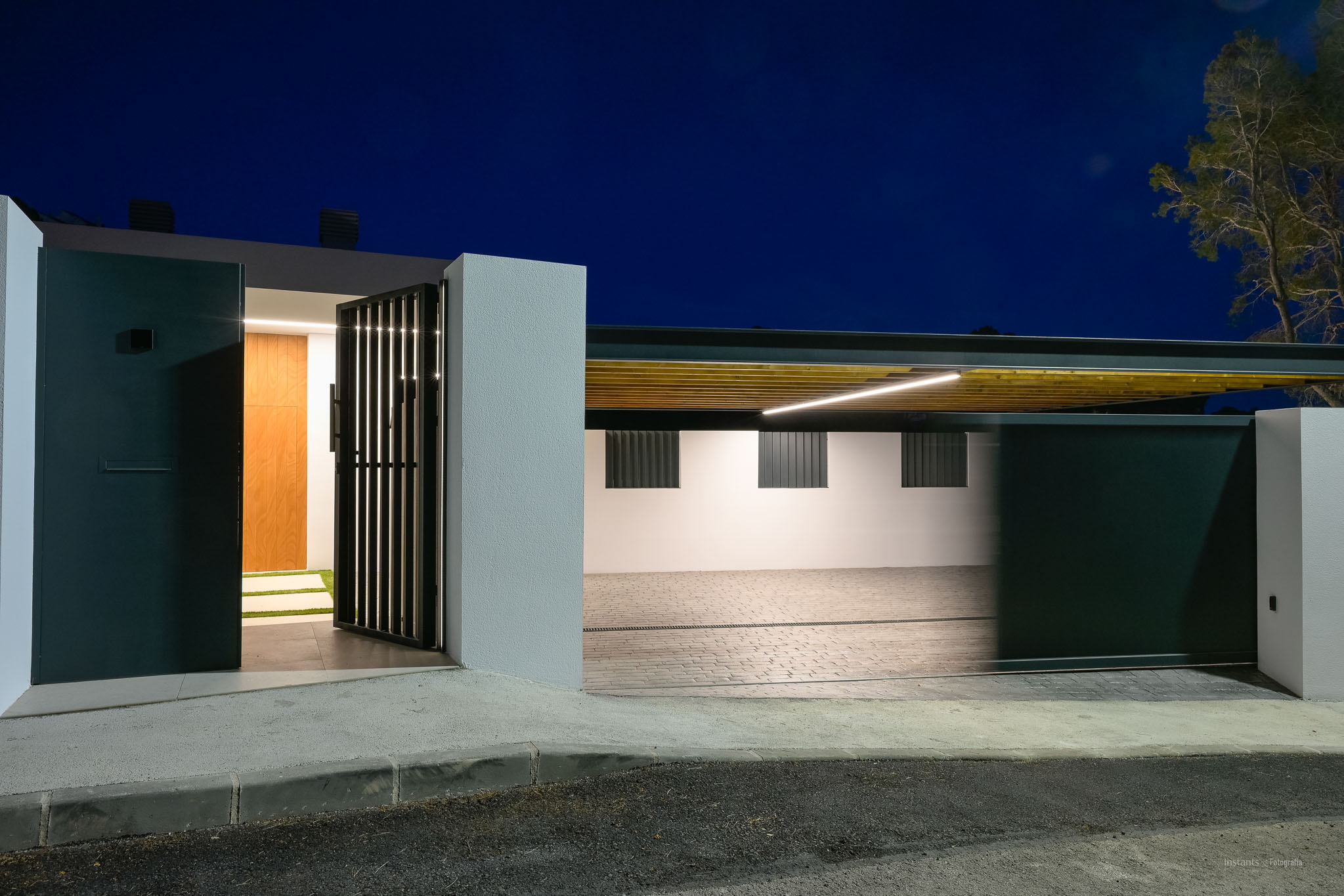 Modern entrance with dark pedestrian gate and wooden main door, alongside an illuminated carport featuring a slatted ceiling and linear LED lighting.