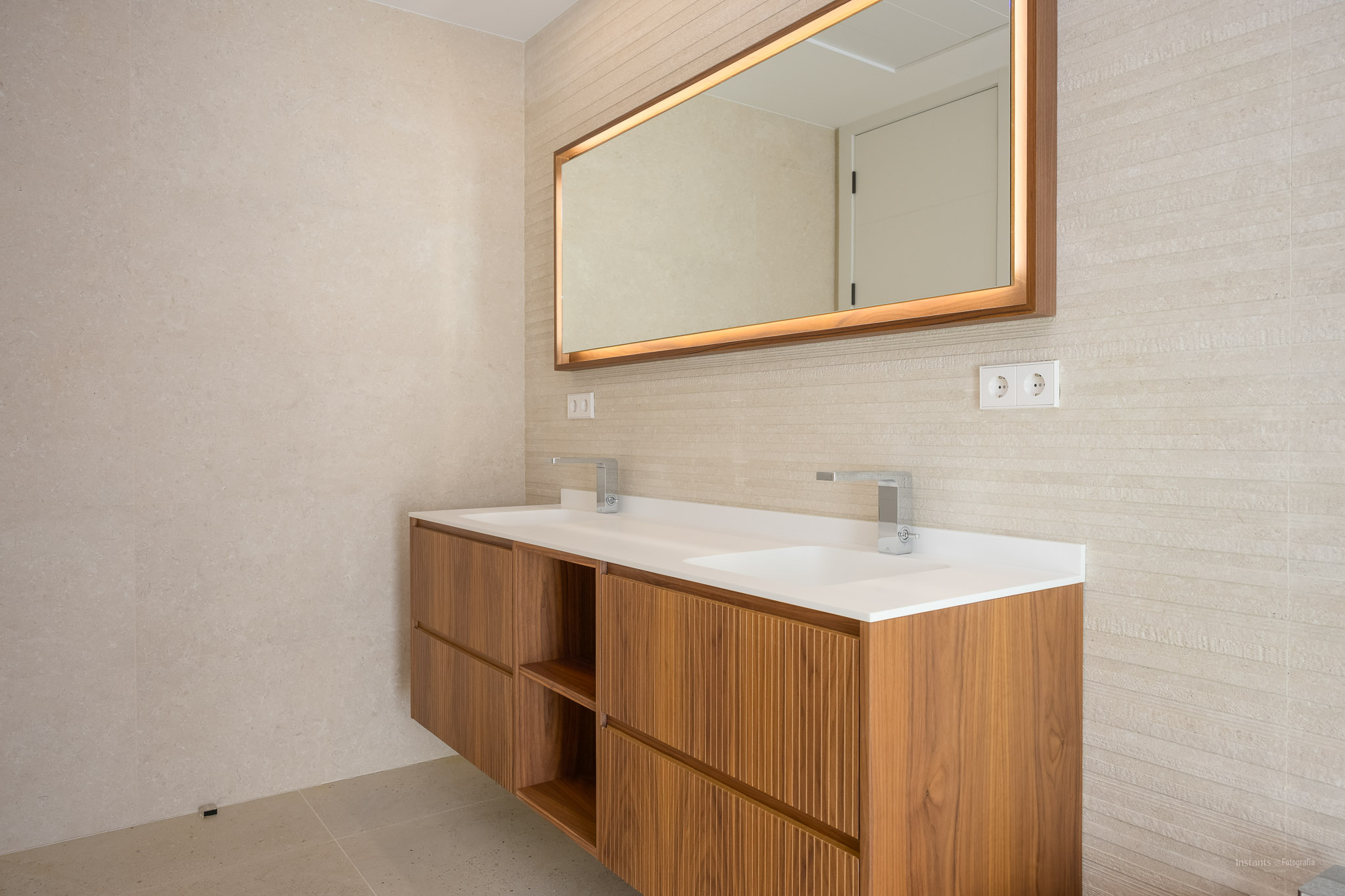 Modern floating wooden bathroom vanity with dual integrated sinks and chrome faucets. Features a backlit mirror with a wooden frame and textured wall tiles.