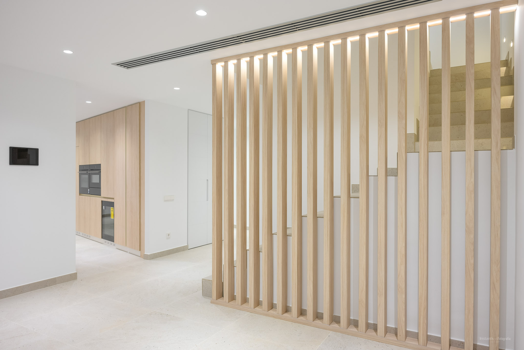 Modern interior featuring a light wood slat partition with integrated lighting, separating a sleek kitchen area with built-in appliances from a contemporary staircase with stone steps.