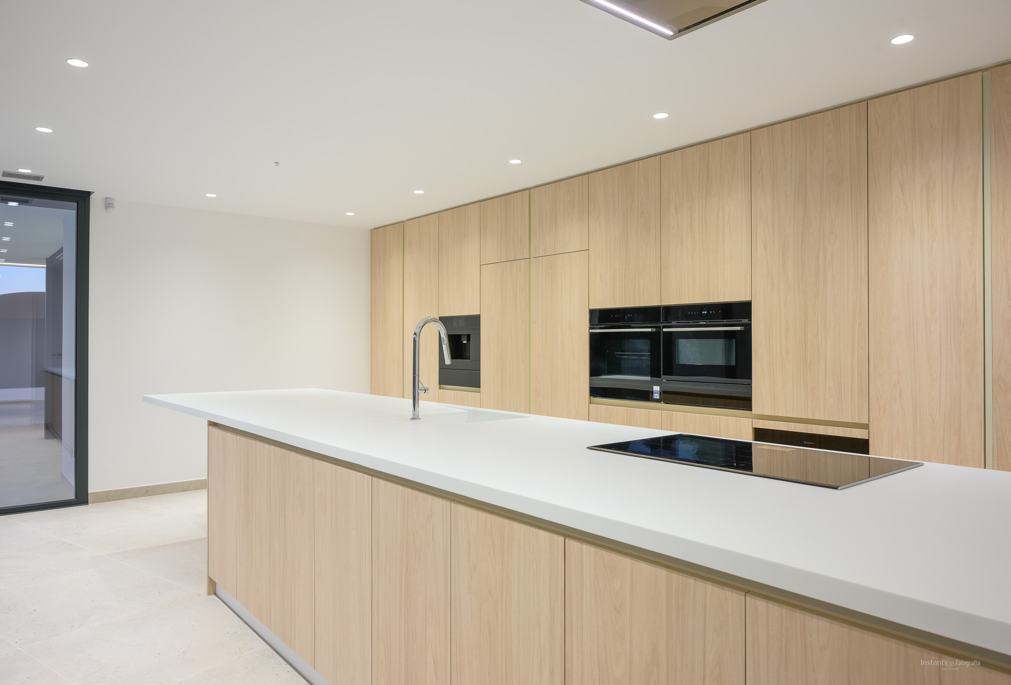 Modern kitchen featuring a large white island with integrated sink and induction hob, complemented by floor-to-ceiling light wood cabinetry and built-in ovens.