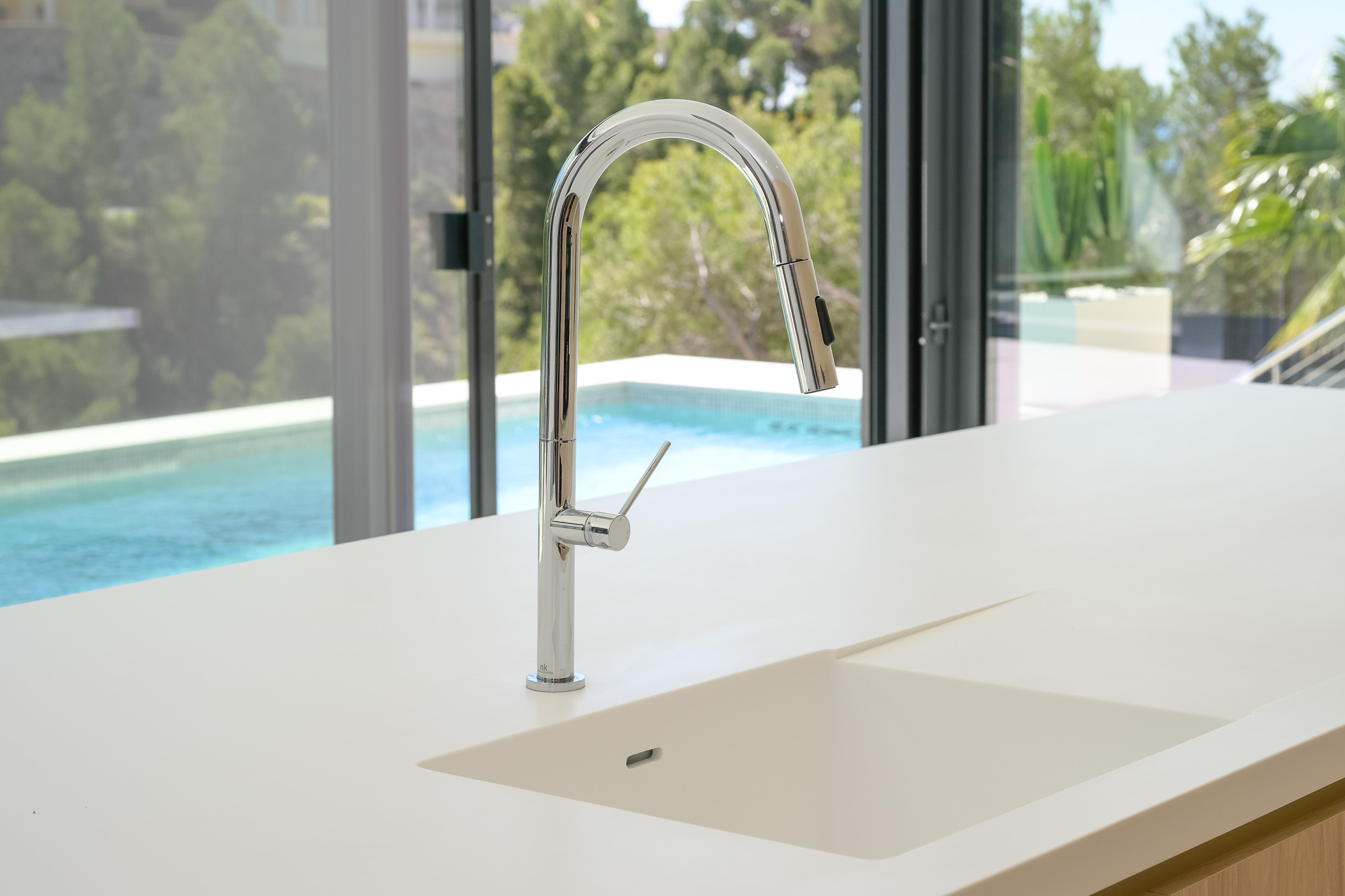 Modern kitchen featuring a light countertop with an integrated sink and a sleek chrome pull-out faucet. Large glass doors offer views of the swimming pool and lush greenery.