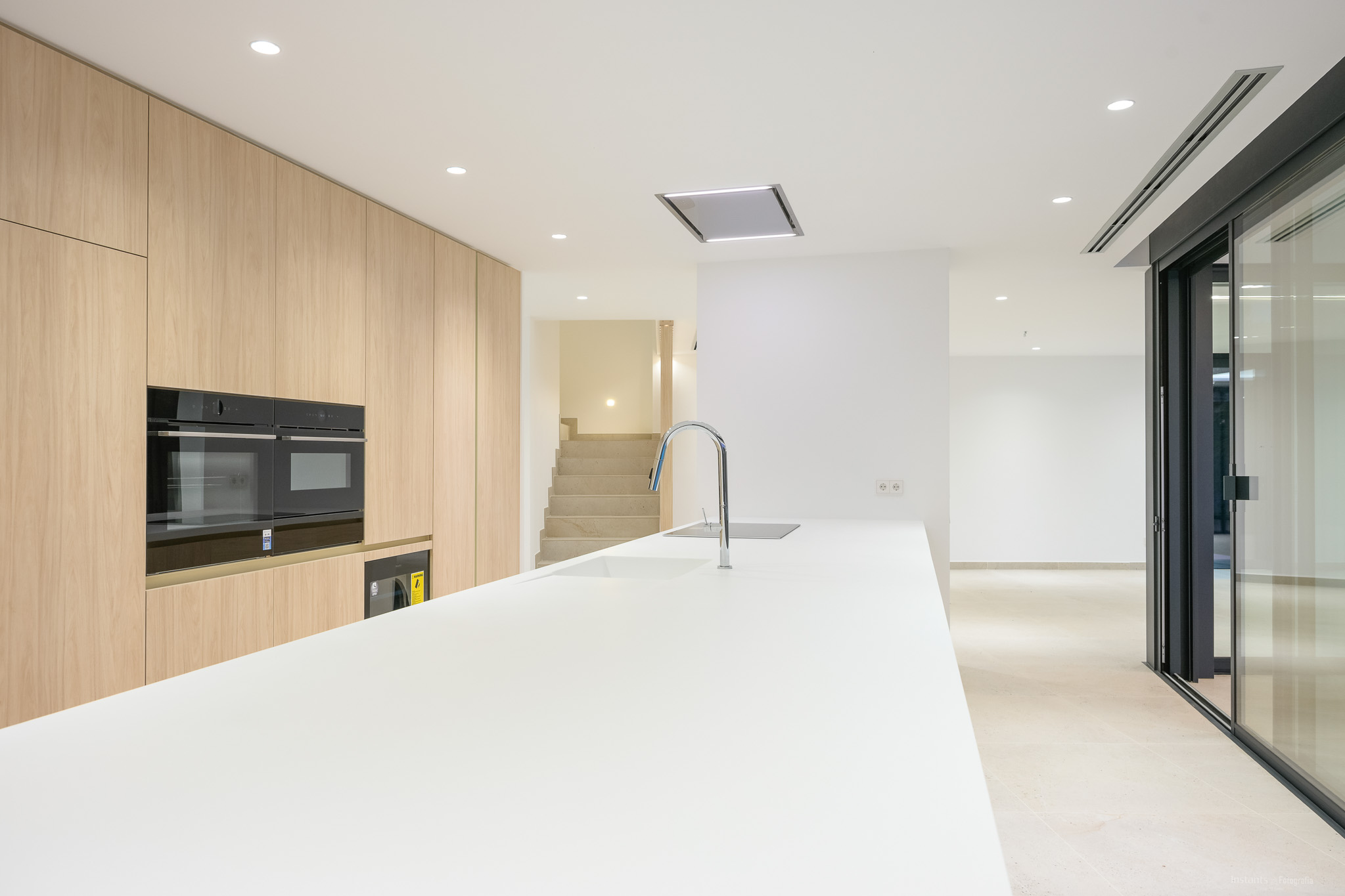 Modern kitchen featuring a long white island with an integrated sink and chrome faucet, complemented by light wood floor-to-ceiling cabinetry and built-in double ovens. Recessed lighting illuminates the contemporary space.