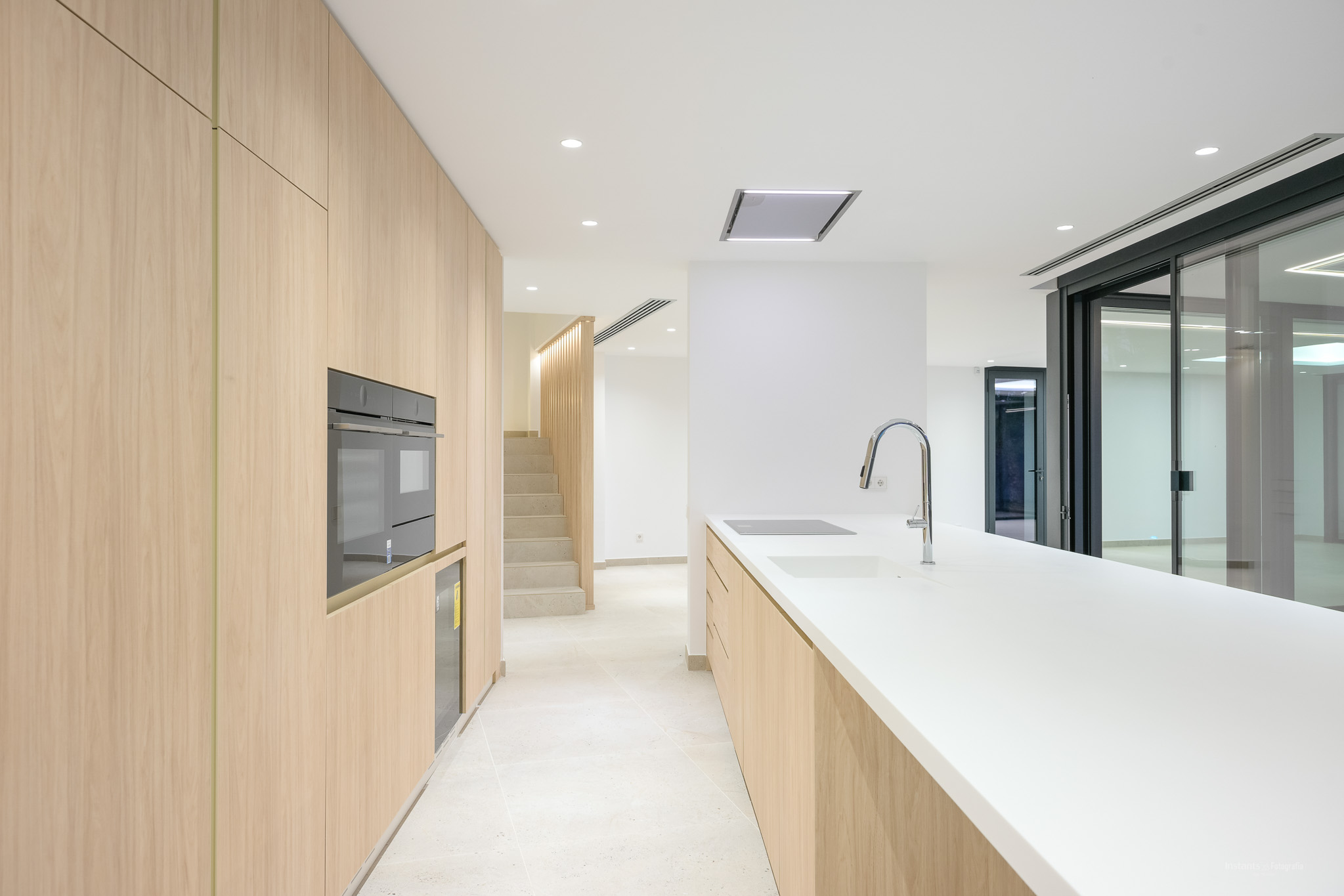 Modern kitchen featuring light wood cabinetry, white countertops, and integrated appliances including an oven, induction hob, and sink with a chrome faucet. Connects to a staircase and outdoor access.