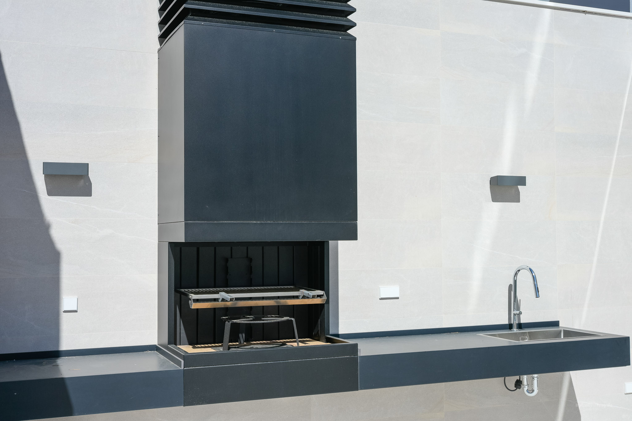 Modern outdoor kitchen featuring a dark integrated barbecue with a sleek hood, a matching counter with a stainless steel sink, and a chrome faucet, set against a light tiled wall.