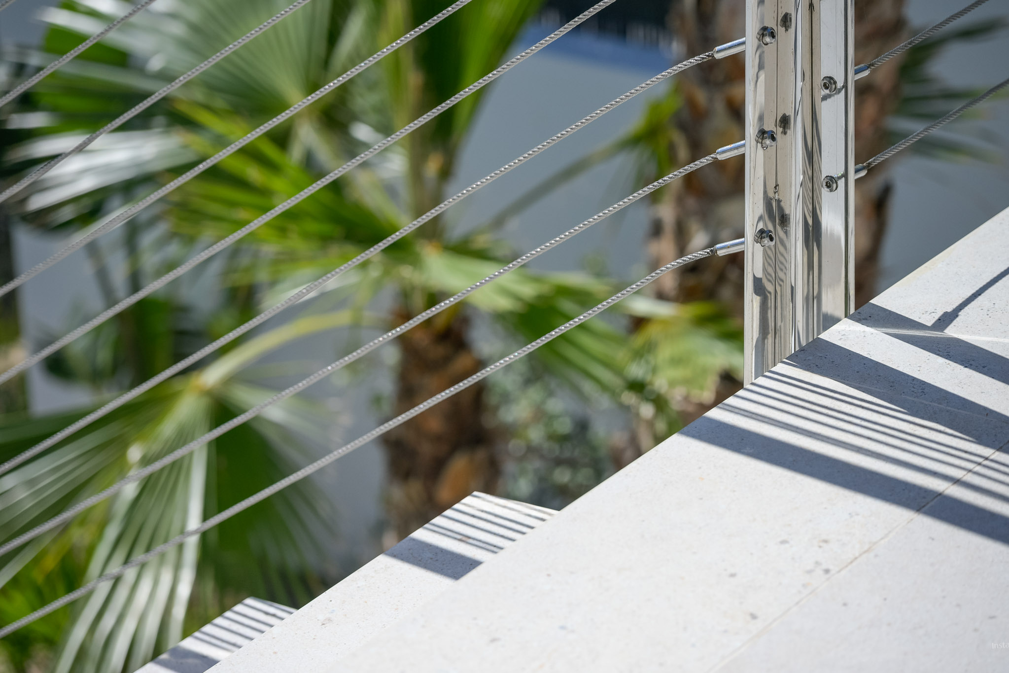 Modern outdoor staircase detail: light-toned steps, polished metal railing with steel cables. Sunlight casts sharp parallel shadows, framed by lush palm foliage.
