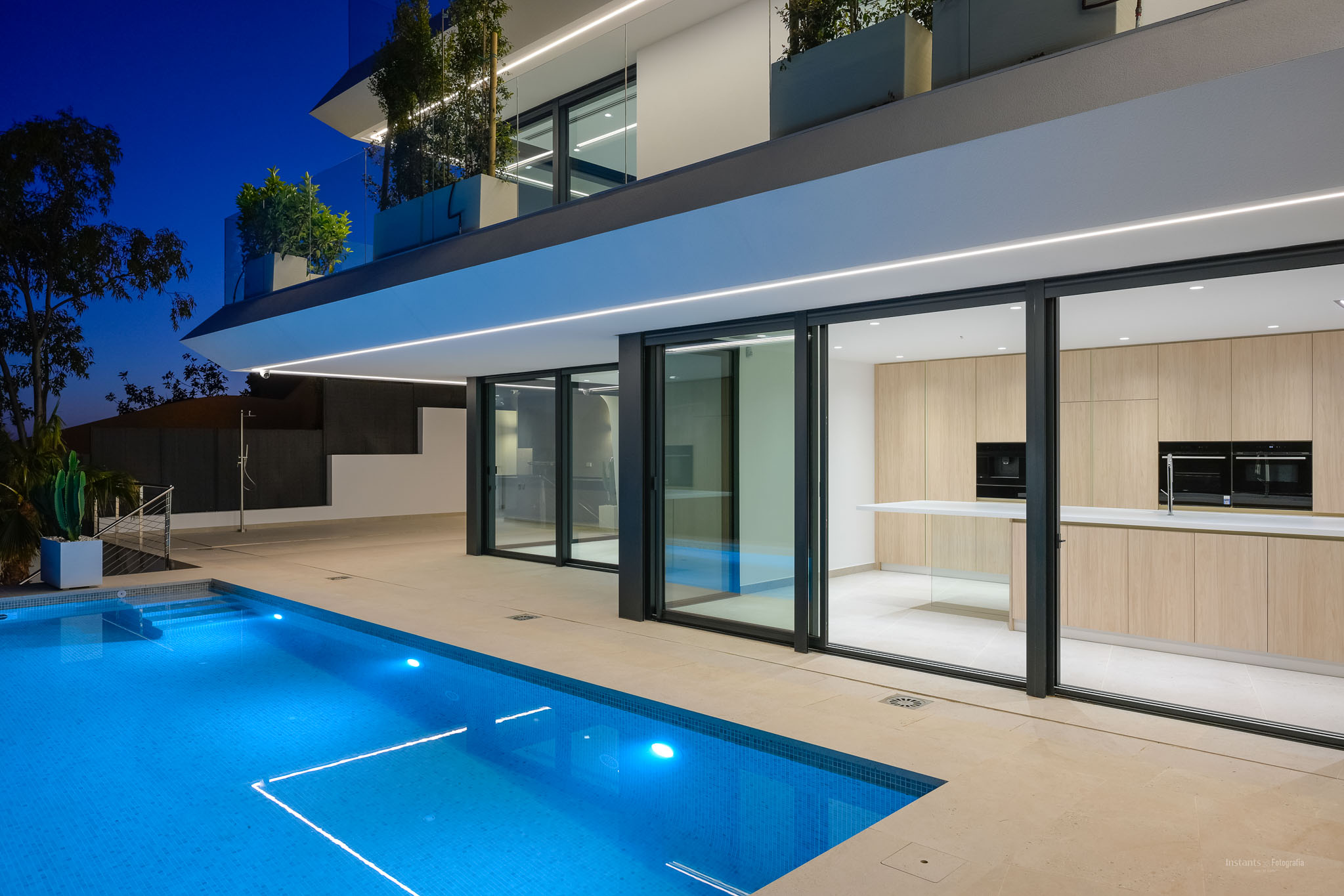 Night view of the modern villa, showcasing the illuminated blue pool and sleek architecture. A light wood outdoor kitchen is visible through the large glass doors.