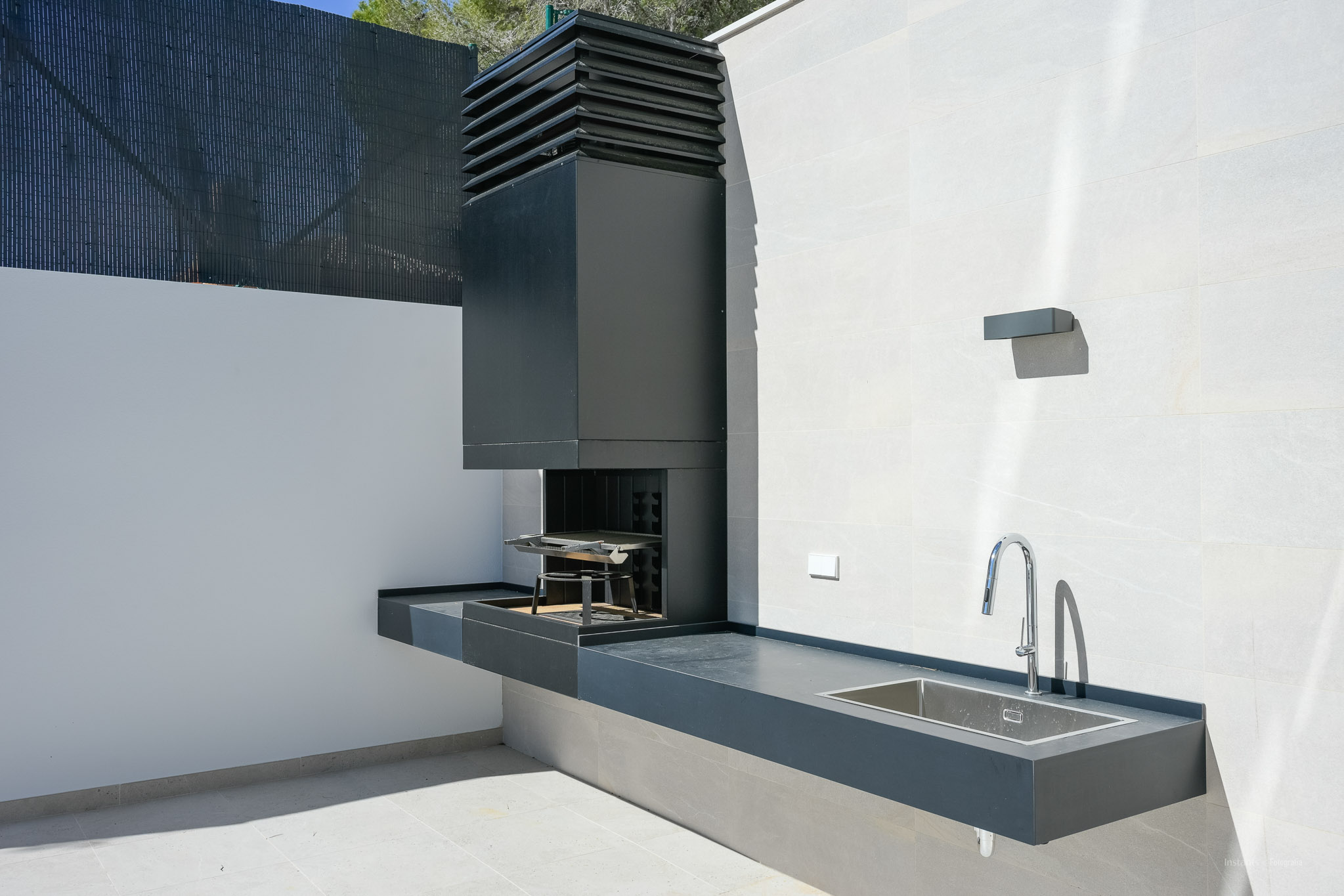 Outdoor kitchen featuring an integrated dark grey BBQ unit, a sleek countertop with a stainless steel sink, and a modern chrome faucet, set against light tiled walls.