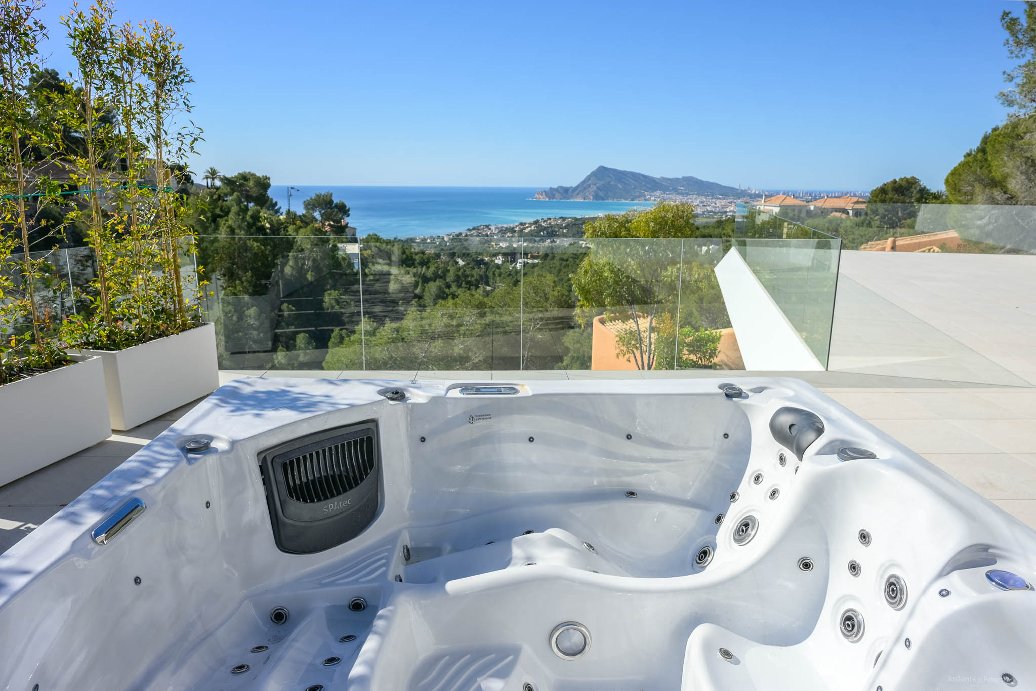 Outdoor spa on a tiled terrace offers panoramic views of the Mediterranean Sea, coastal town, and distant mountains. Glass railings ensure an unobstructed vista, complemented by lush greenery.