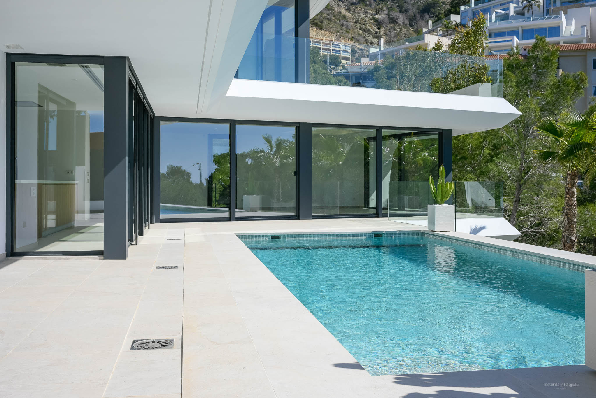Private swimming pool with blue mosaic tiles and light stone terrace, bordered by large glass doors connecting to the villa's modern interior.