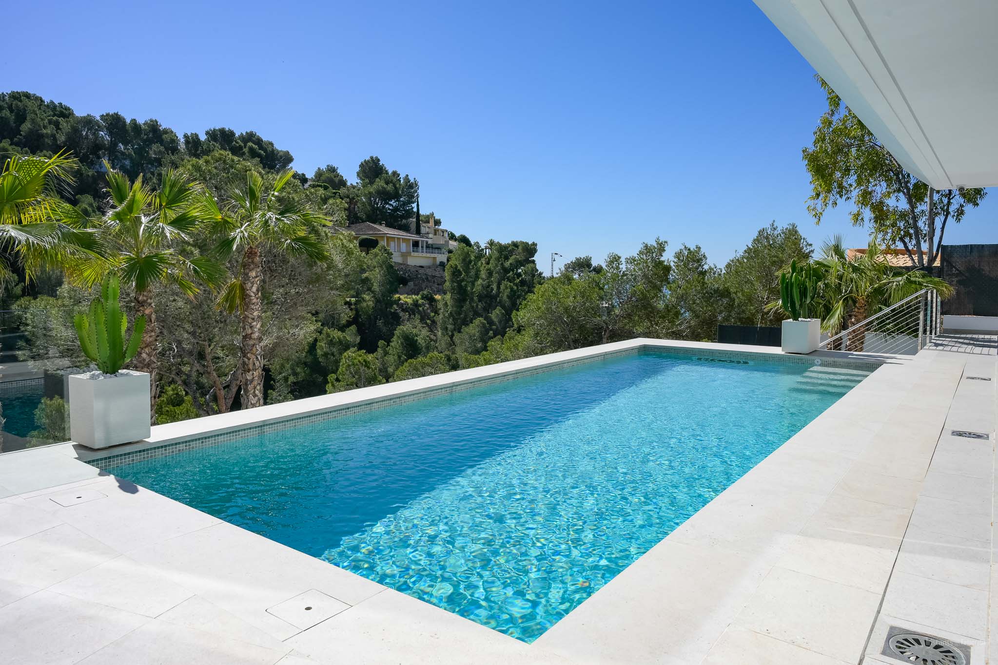 Rectangular swimming pool with light blue mosaic tiles and white stone coping, set against lush Mediterranean greenery under a clear blue sky.