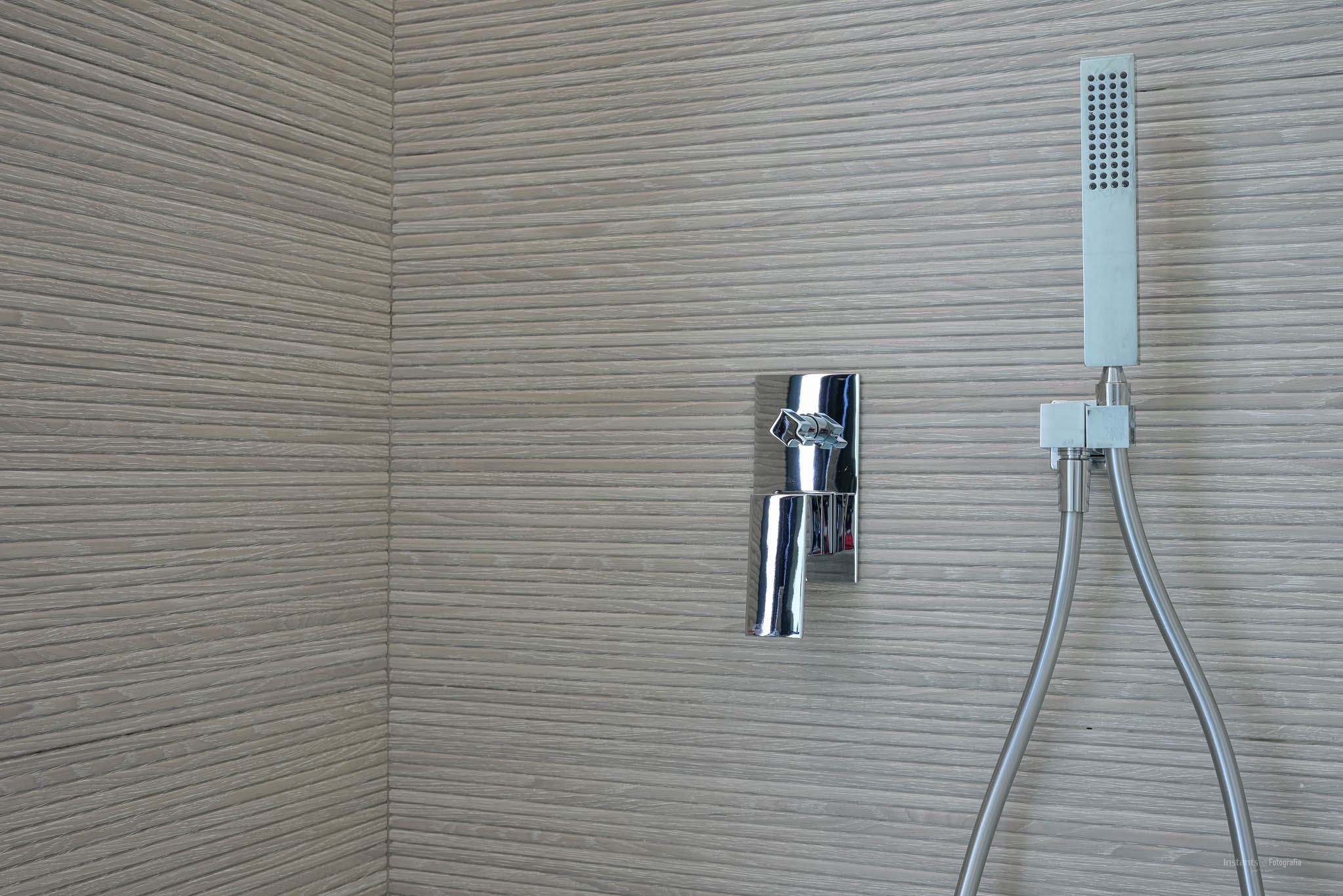 Shower area featuring chrome mixer and handheld shower against a wall clad with horizontally grooved textured tiles.