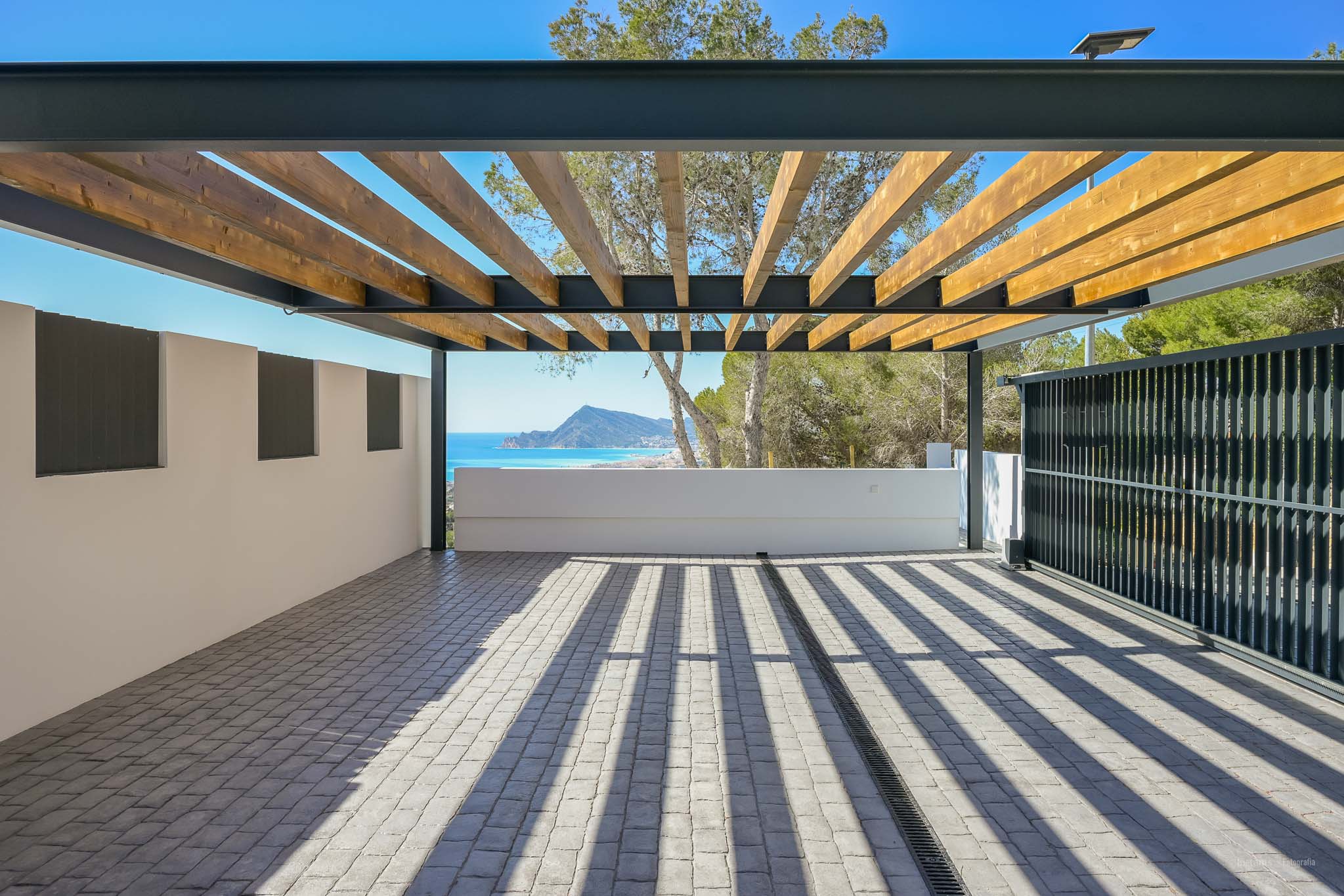 Spacious outdoor area featuring a pergola with wooden beams and a metal frame over cobblestone paving. Enjoy panoramic sea and mountain views, with natural light casting distinct shadows.
