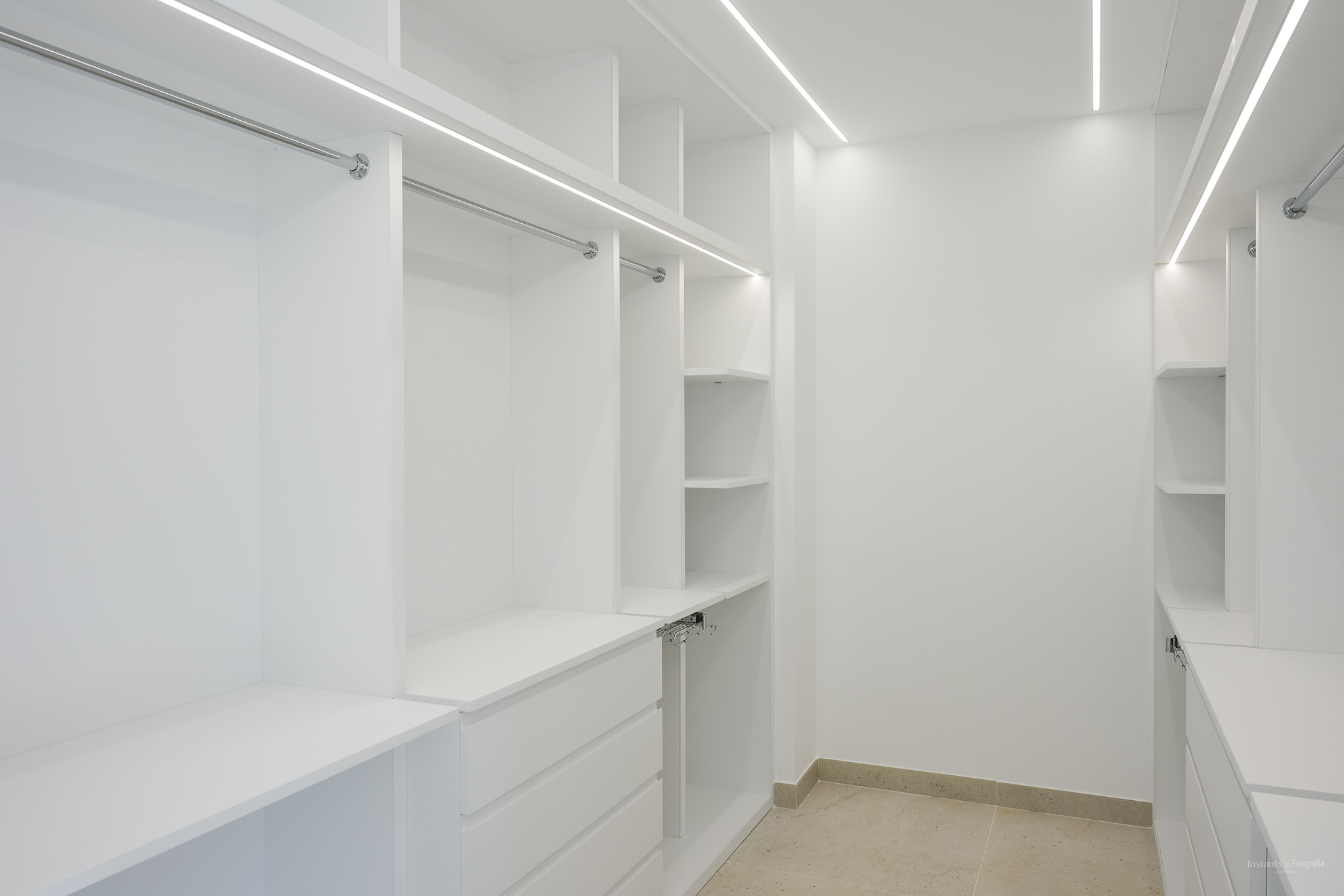 Spacious walk-in closet featuring custom white cabinetry, chrome hanging rails, and integrated LED strip lighting for optimal illumination. Includes multiple shelves and drawers, set on light-toned tiled flooring.