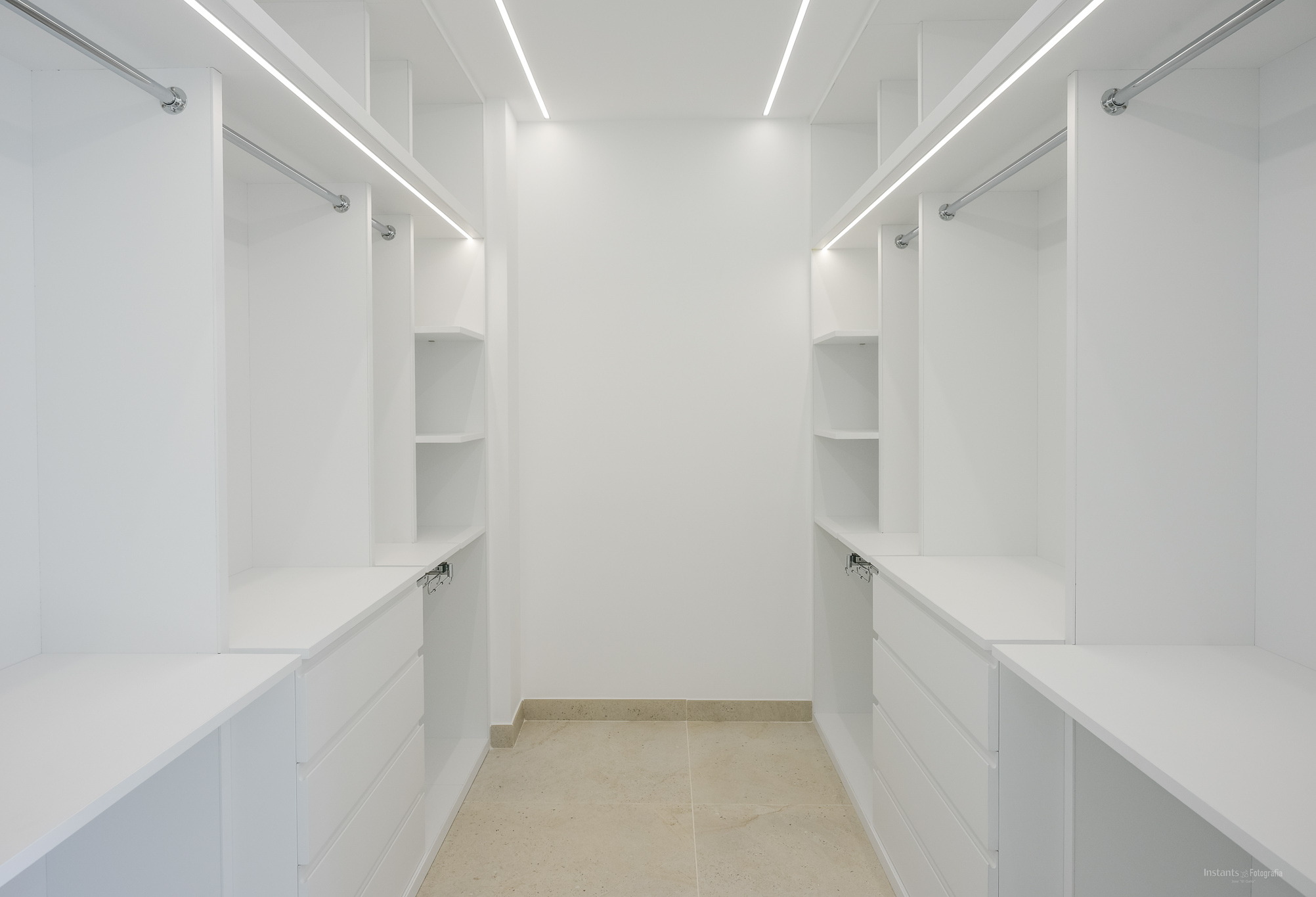 Spacious white walk-in closet featuring custom shelving, drawers, and hanging rails, illuminated by integrated LED strips and light tiled flooring.