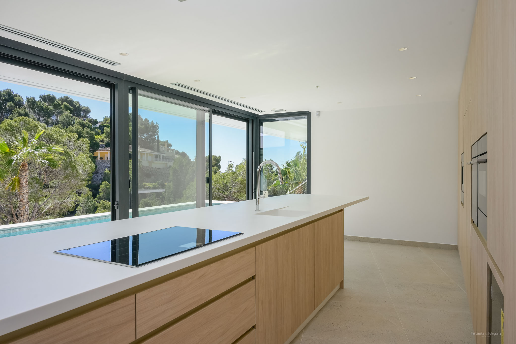 The modern kitchen features a large island with a white countertop, integrated induction hob, and chrome faucet. Sliding glass doors offer views of lush Mediterranean greenery and a private pool.