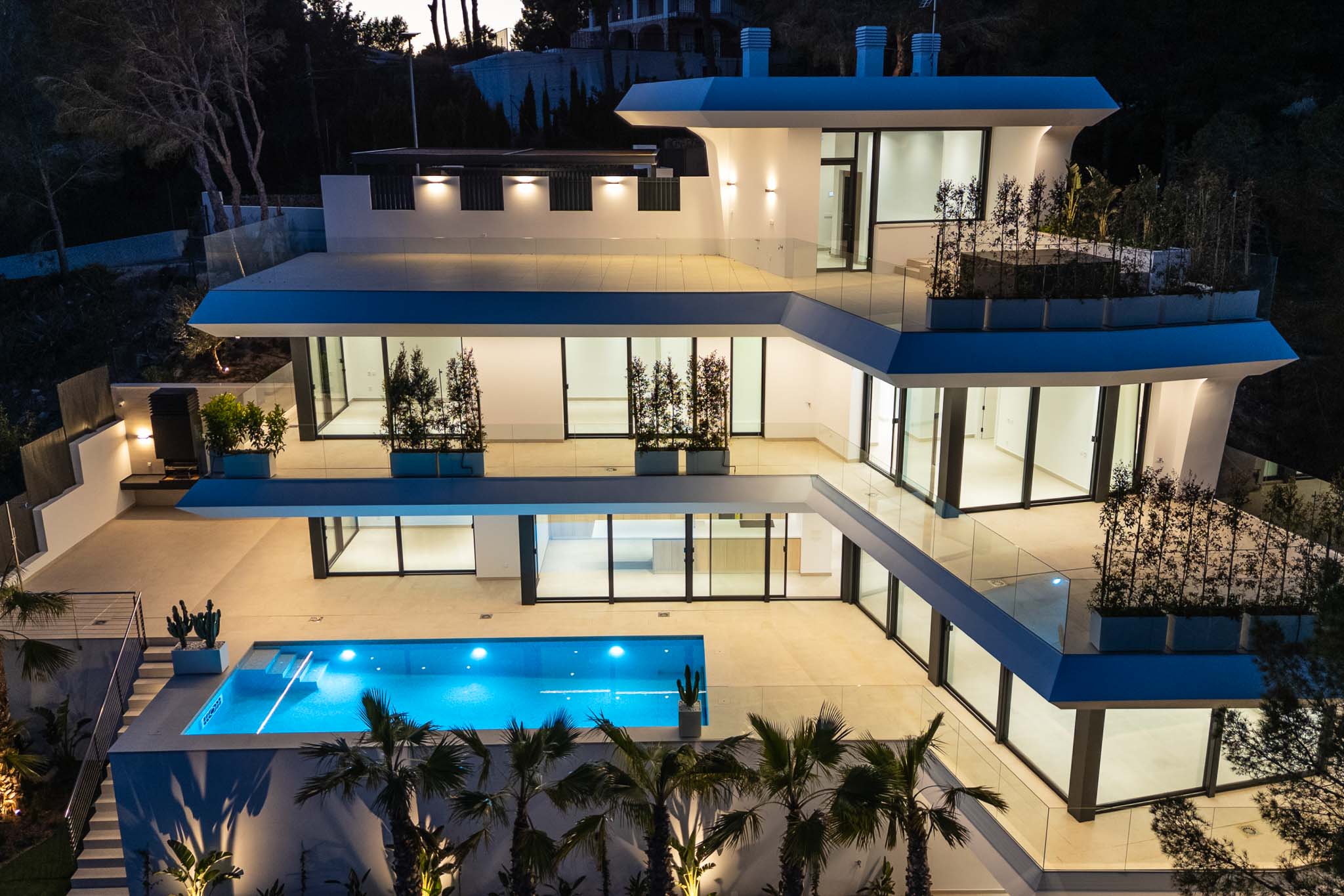 The modern villa's multi-level architecture is illuminated at dusk, showcasing its vibrant blue pool and expansive terraces with glass railings.