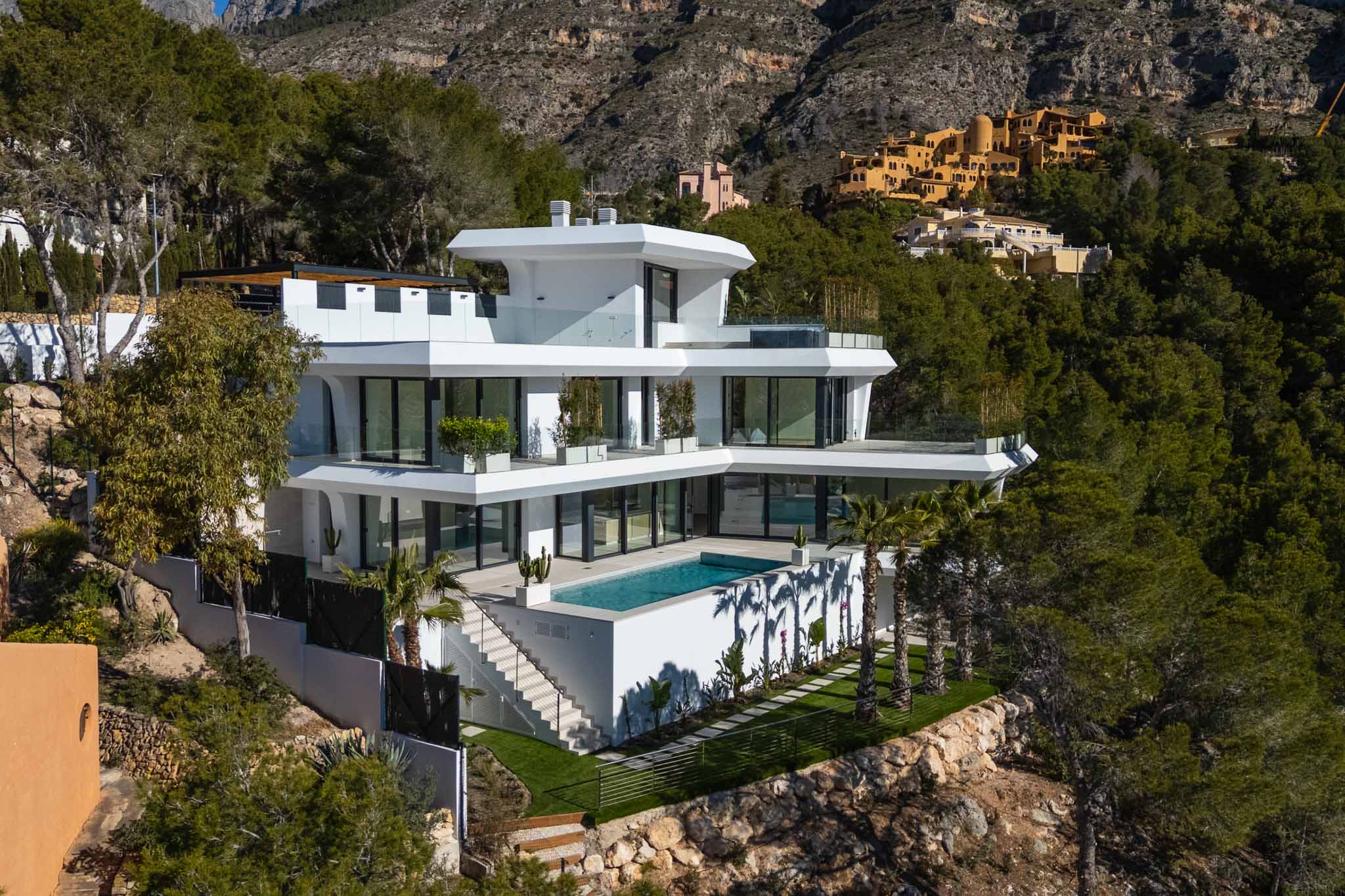 This multi-level contemporary villa features a pristine rectangular pool and expansive terraces with glass railings, nestled into a verdant hillside with mountain views.