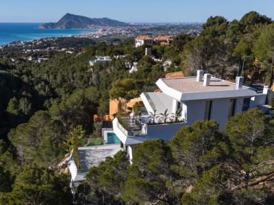 Aerial view of a modern white villa cascading down a pine-covered hillside, offering expansive panoramic views of the Mediterranean Sea and Altea's coastline. A private pool is visible on a lower terrace.