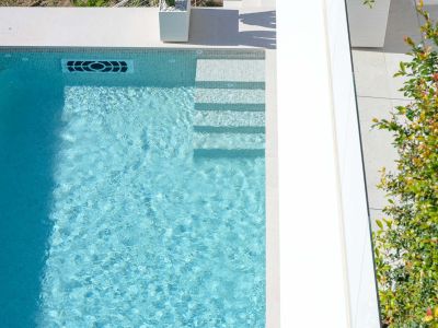 Aerial view of the light blue mosaic tiled pool with integrated steps, featuring a modern water return system and surrounding light-colored paving.