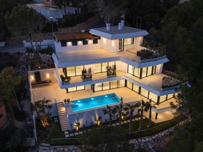 Aerial view of the modern multi-level villa at dusk, showcasing its illuminated terraces, bright blue swimming pool, and an outdoor kitchen area with elegant lighting amidst lush greenery.