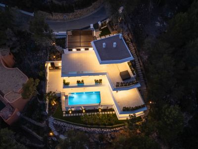 Aerial view of the modern villa at night, showcasing its brightly lit terraces and the inviting, glowing blue swimming pool. Multiple outdoor living areas are visible, set against the dark, natural hillside.