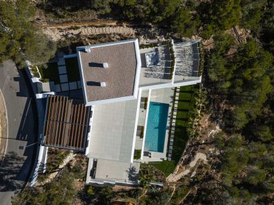 Aerial view showcasing the modern villa's architecture, including a rectangular swimming pool, expansive paved terraces, and an outdoor spa, all nestled within natural Mediterranean greenery.
