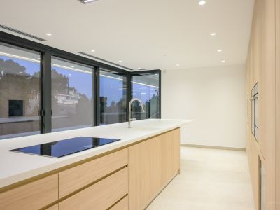 Contemporary kitchen featuring a large central island with a white countertop, integrated induction hob, and chrome faucet. Light wood cabinetry and built-in ovens line the wall, complemented by expansive windows.