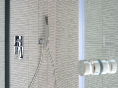 Detail of a contemporary shower featuring grey textured rectangular tiles, a sleek chrome mixer tap, and a flexible handheld showerhead. A glass screen is partially visible.