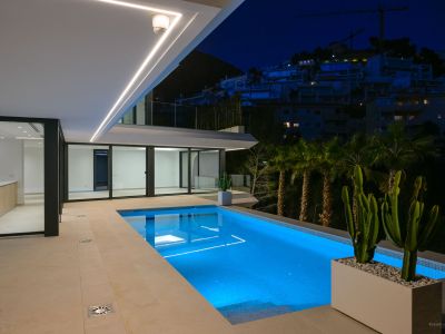 Illuminated swimming pool at night, featuring vibrant blue underwater lighting and a spacious tiled terrace. Modern linear ceiling lights enhance the sleek architecture, overlooking a hillside with distant buildings.