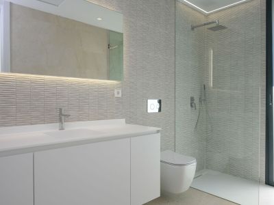 Modern bathroom featuring a sleek white vanity with integrated sink, backlit mirror, and textured grey wall tiles. A wall-mounted toilet and spacious walk-in shower with glass partition.