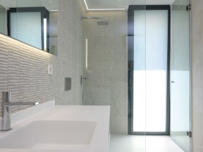 Modern bathroom featuring a white basin, chrome faucet, and textured grey wall tiles. Includes a glass-enclosed shower with a rainfall head and LED-lit mirror.