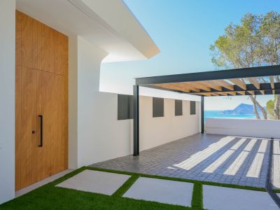 Modern entrance featuring a tall wooden door, artificial grass path, and a paved parking area with a metal pergola. Distant Mediterranean sea and mountain views are visible.